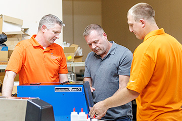 Three men in orange and gray shirts stand around a blue machine in a workshop or warehouse, examining and discussing the equipment.