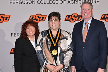 Angel Riggs stands in the middle of the photo, wearing a medal. Jayson Lusk, vice president and dean of OSU Agriculture, stands to the right, wearing a dark suit. Cynda Clary, associate dean of academic programs, stands to the left. They stand in