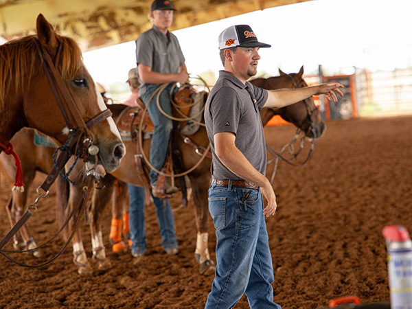Joby Bush, in a gray polo shirt, jeans and a baseball cap points across an indoor rodeo arena while several riders on horses wait behind him on the dirt floor.