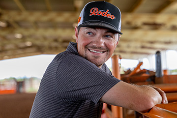 A smiling man in a black rodeo cap and dark shirt leans on a fence inside a rodeo arena.