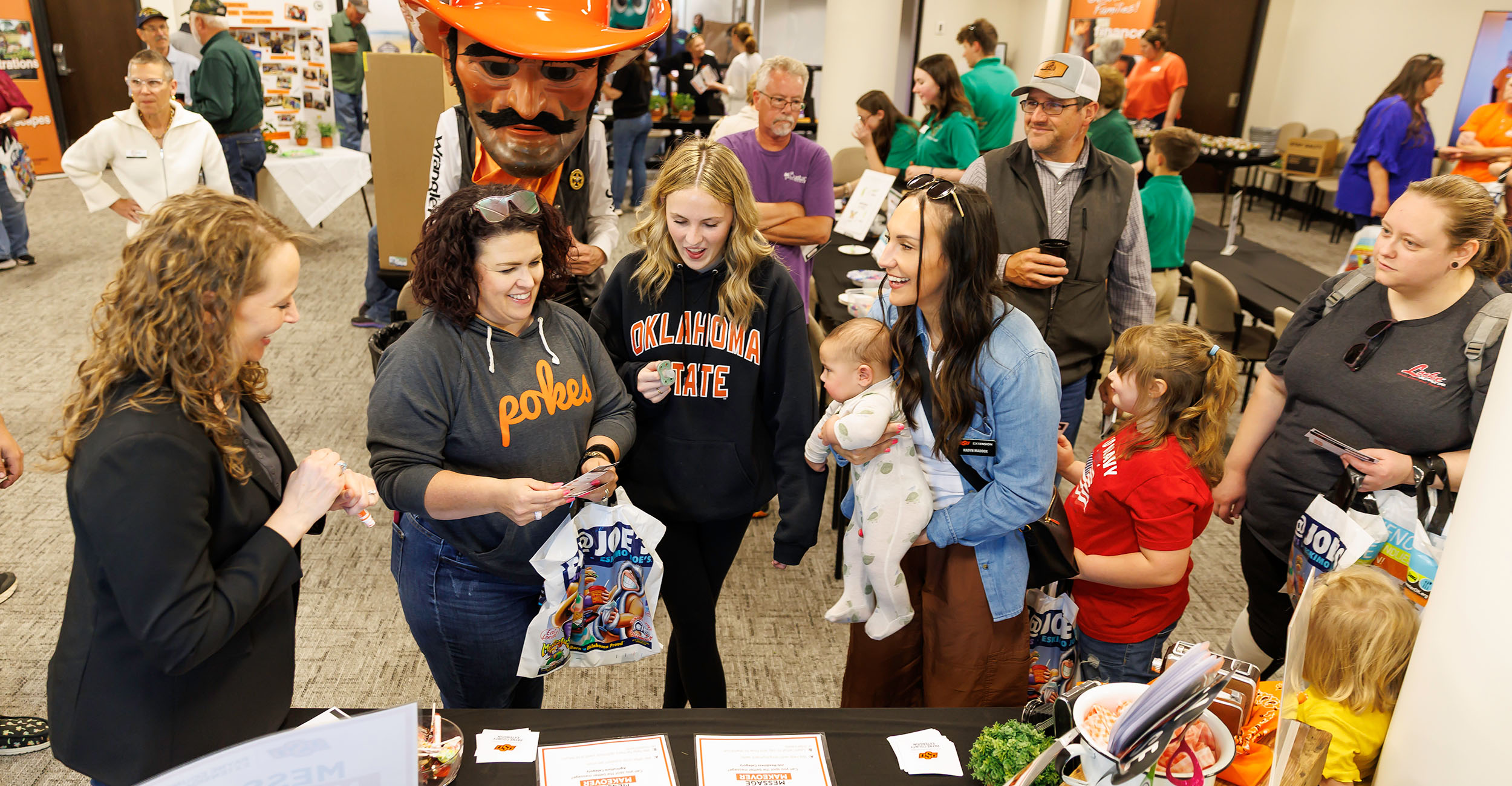 OSU Extension educators stand around a display table, showing off programming to patrons. The group is inside the Payne County OSU Extension office. Pistol Pete stands in the background. 