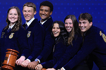 Six smiling teenagers wearing navy blue FFA jackets with embroidered gold emblems pose together against a blue background, placing their hands on a wooden gavel.