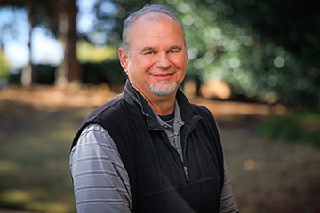 Sam Fuhlendorf, with short gray hair and a goatee, smiles and stands outdoors in a wooded setting, wearing a dark fleece vest over a striped polo shirt.