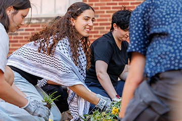 OSU students, including Sereen Elkhalid, plant flowers in the Heritage Garden outside a brick campus building. Elkhalid wears gardening gloves and a black-and-white patterned shawl as she works alongside fellow volunteers planting native greenery.