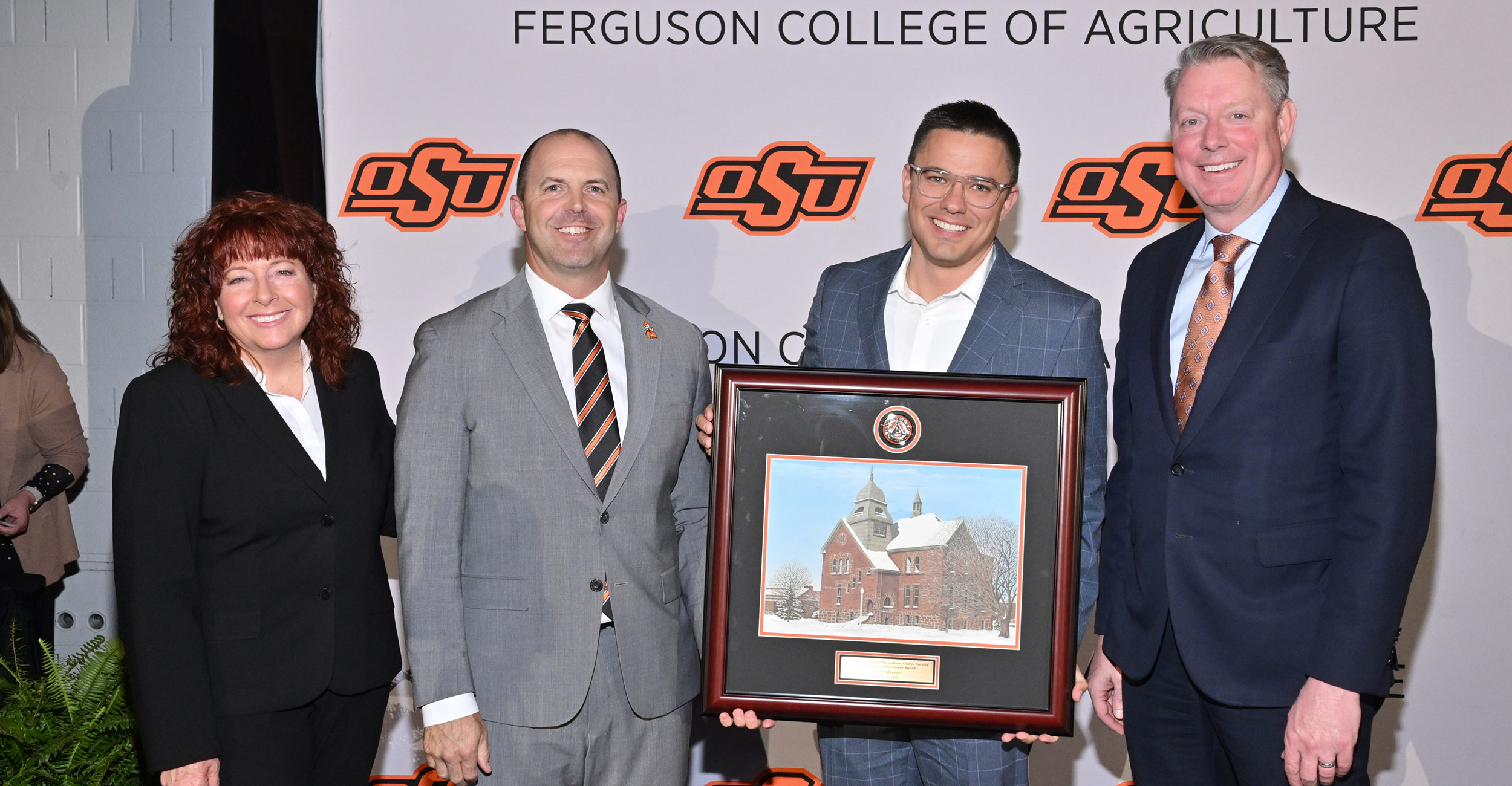 Jeremy Bennett wears a gray suit and stands in the middle of the frame. He holds a framed picture of Old Central. Cynda Clary, associate dean of the Ferguson College, Justin Anderson, president of the Ferguson College Alumni Society, and Jayson Lusk, vice president and dean of OSU Agriculture, stand around Bennett. 