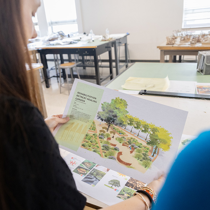 Izzy Holding stands in the foreground, holding a printing of her healing garden design. Professor Qing Luo stands in the foreground. 