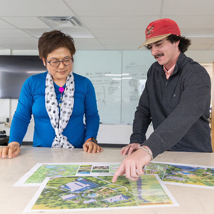 Carson Holmes stands to the right and points to a piece of paper on a table. Professor Qing Luo stands to the left and listens to him.