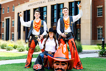 Hunter Morton stands to the left, wearing his Pistol Pete outfit and holding his finger in a pistol form. Pete Littlefield stands to the right doing the same pose. Olivia Klug kneels in the center next to the Pistol Pete headgear. The group faces