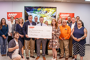 Brian Whitacre stands in the middle of the photo, holding up a novelty check. He is surrounded by members of OSU Extension, AARP Oklahoma and the Fairview Public Library.