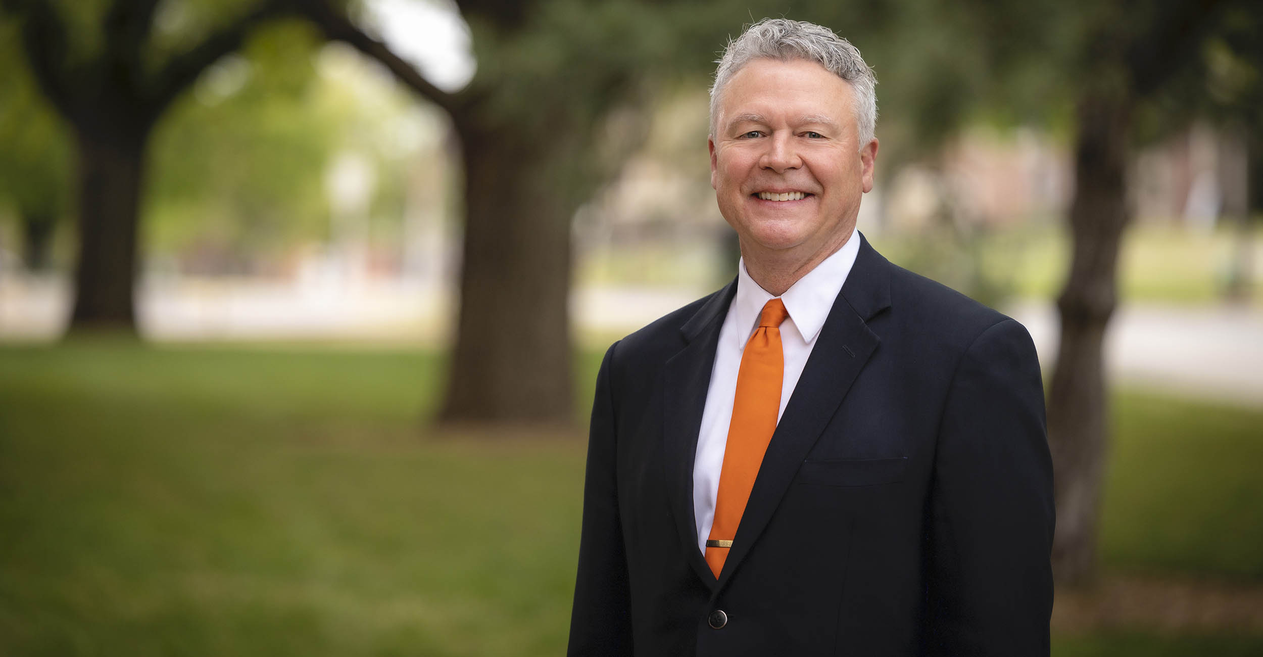A person in a dark suit with an orange tie stands outdoors on a tree-lined campus lawn, posed for a professional portrait with blurred green foliage and a walkway in the background.