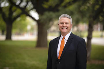 A person in a dark suit with an orange tie stands outdoors on a tree-lined campus lawn, posed for a professional portrait with blurred green foliage and a walkway in the background.