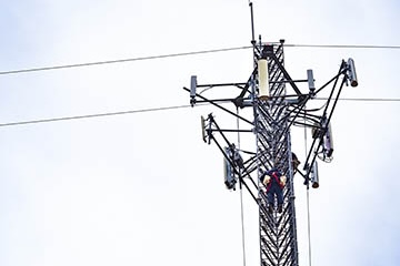 Worker climbs a tall telecommunications tower fitted with antennas and equipment, illustrating broadband infrastructure in a rural or utility setting.