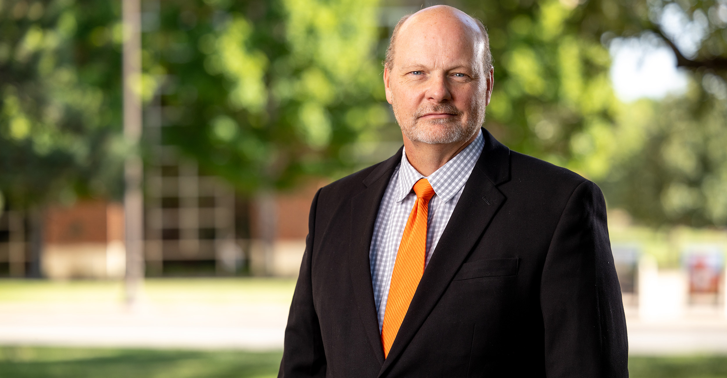 A professional outdoor portrait shows a person in a dark suit with a bright orange tie standing on a campus lawn with trees and blurred academic buildings in the background.