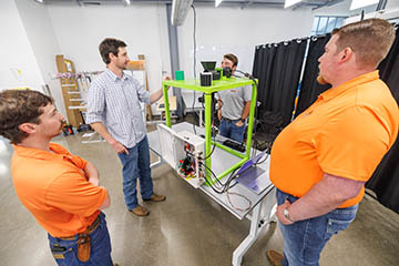 Four people stand around a worktable in a laboratory, examining and discussing a bright green-framed technical device connected to wires, sensors, and a laptop, indicating a hands-on engineering project or demonstration.