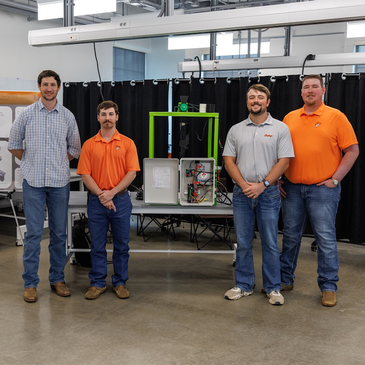Four people stand in a lab space posing beside a green-framed engineering device with an open control panel and visible wiring, presenting a completed technical project setup.