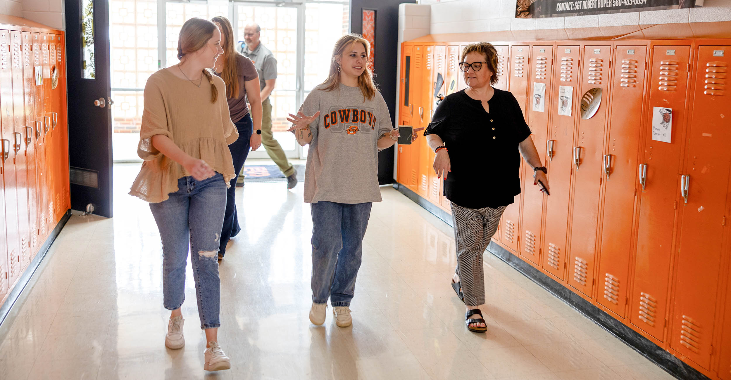 Three people walk and talk down a school hallway lined with bright orange lockers while a few others move in the background near the entrance.