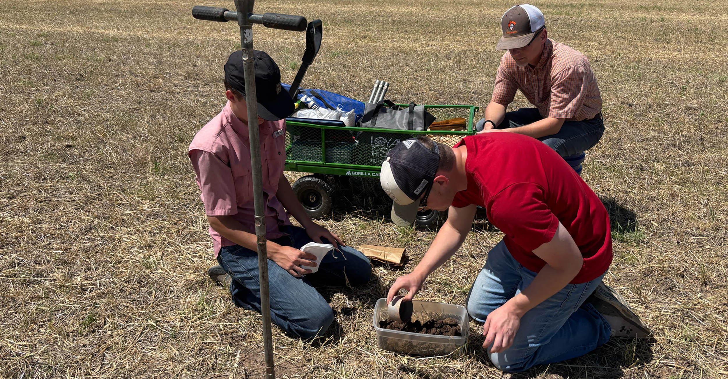 Three students kneel in a dry grassy field collecting and examining soil samples using handheld tools and containers beside a small utility cart and a vertical probe.