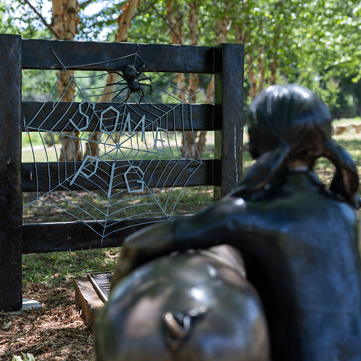 A metal sign featuring the words “Some Pig” inside a spider web with a spider sculpture is mounted on a fence, while a bronze statue of a child looks on, referencing a scene from Charlotte’s Web along a wooded storybook trail.