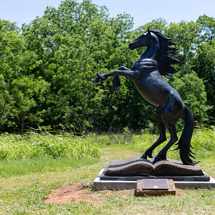 A bronze statue of a rearing horse standing atop an open book is displayed outdoors in a grassy clearing with trees in the background, symbolizing a literary-themed trail installation.