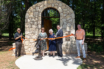 A group of five people stand in front of a stone archway cutting an orange ribbon beside a bronze statue, marking a ceremonial opening or dedication of an outdoor site.