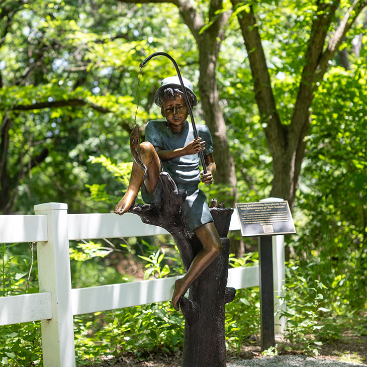 A bronze statue of a child sitting on a tree stump and holding a fishing pole stands beside a white fence and informational sign along a shaded wooded trail.
