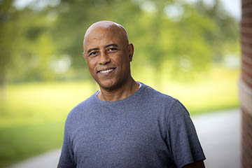 A close-up outdoor portrait shows a person in a gray shirt standing in front of a softly blurred green landscape, with a brick wall visible along the edge of the frame.