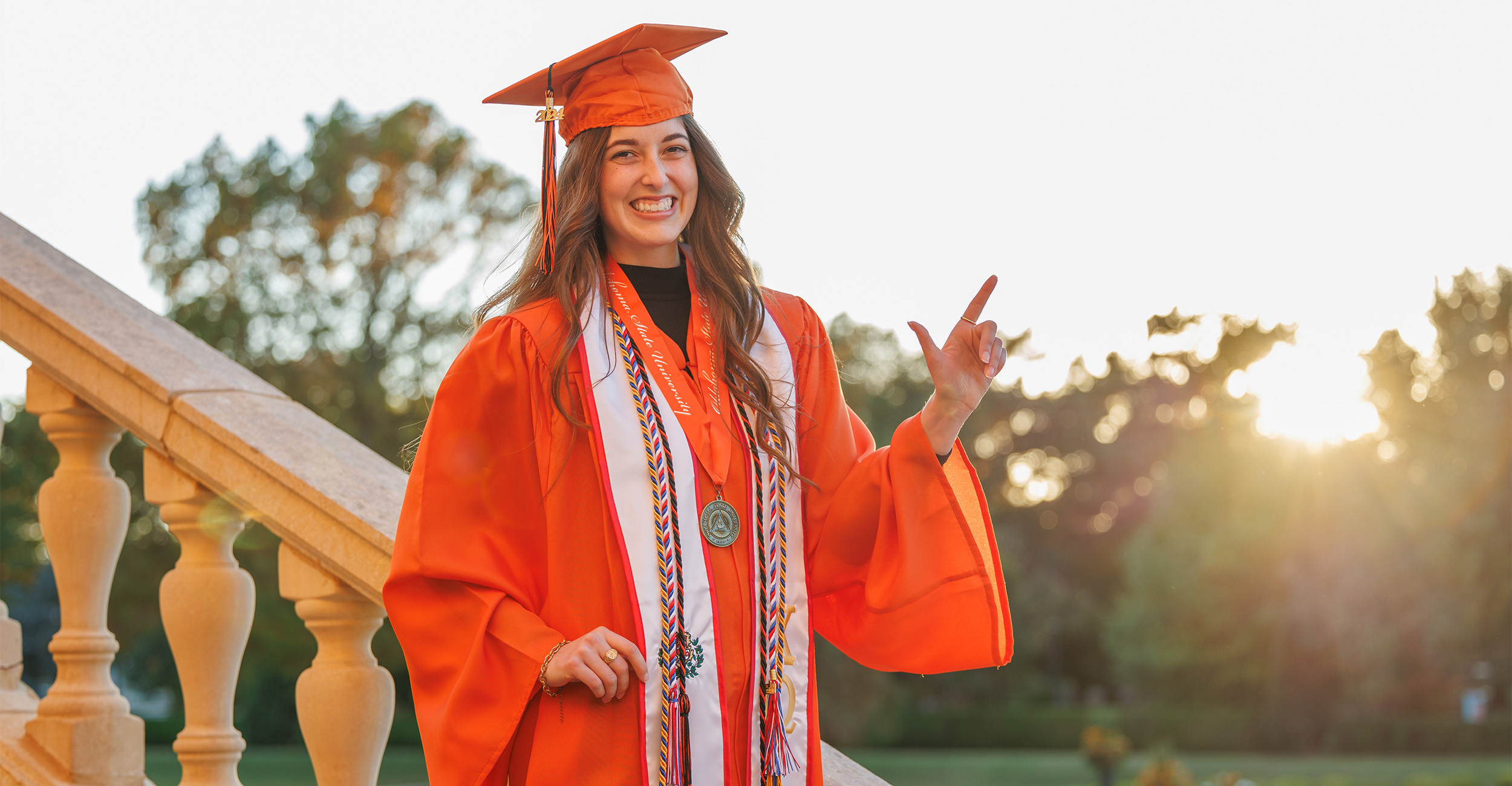 Photo of Ferguson College student Karstyn Cantrell wearing an orange graduation cap and gown with regalia from various student organizations. She has long, curled brown hair, an enthusiastic smile and is standing on a staircase at Oklahoma State University and holding up a "pistol's firing" hand sign.