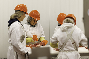 Four people wearing white protective coats, orange hard hats, hairnets, and gloves stand around a processing table in a clean industrial facility, handling cuts of meat during a hands-on training or production activity. White paneled walls and