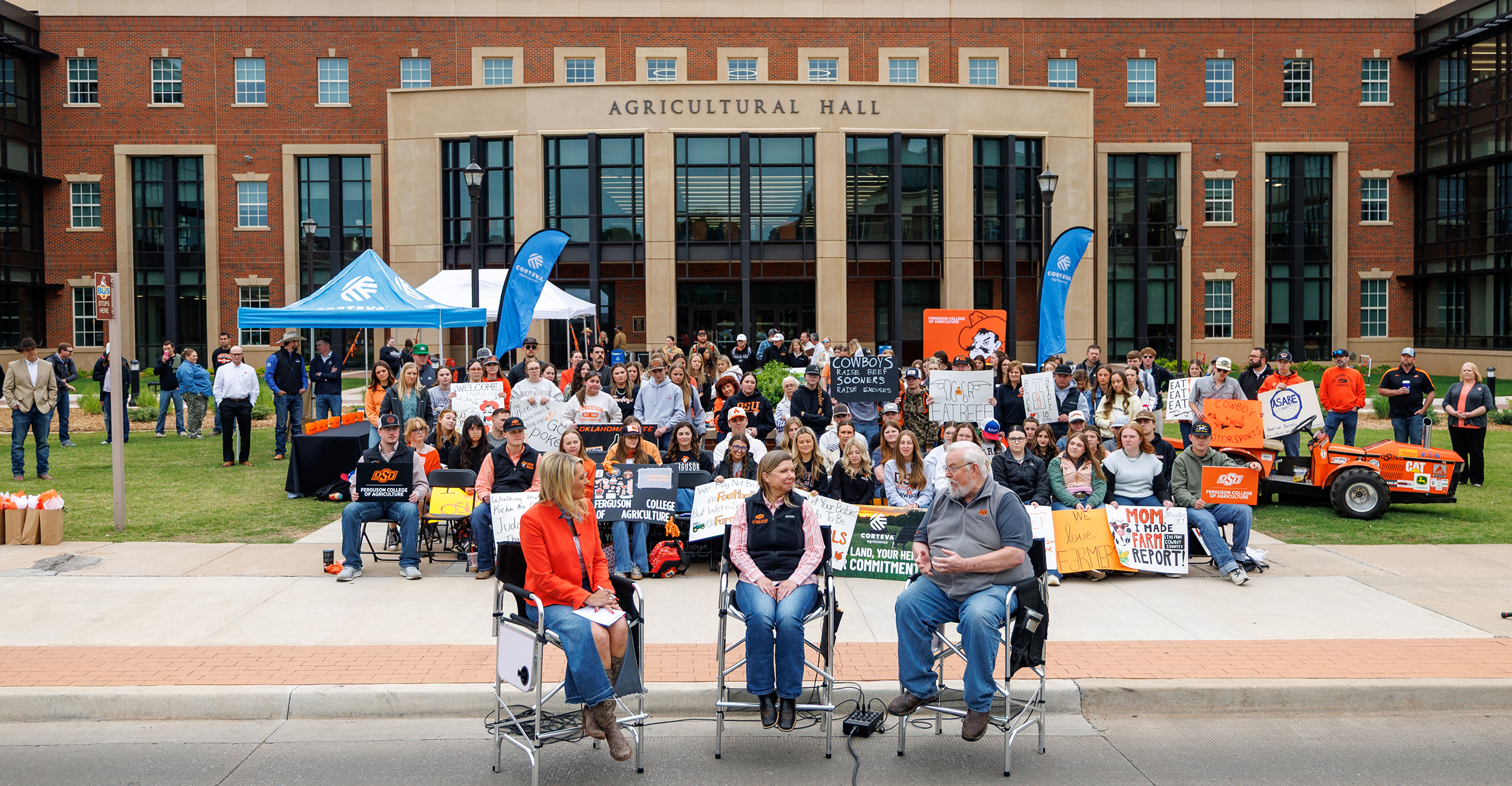 A large group of people gathers outdoors in front of a brick building labeled “Agricultural Hall,” sitting and standing together for a group photo behind two seated speakers with microphones and event displays in the background.