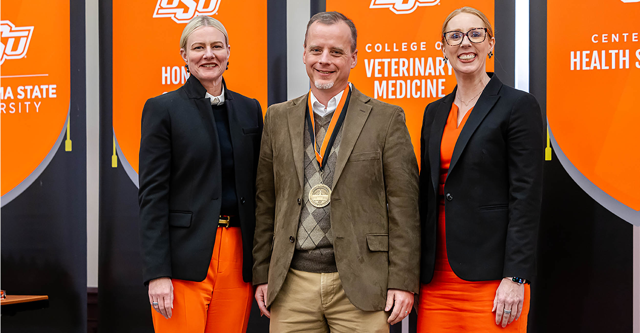 Left to right is Kayse Shrum wearing a black sweater and blazer with orange pants. Her blonde hair is slicked back. To her right is Tyson Ochsner in khaki pants, a tan sweater and a tan sport coat with an OSU medal hanging from his neck. On the far right is Jeanette Mendez in glasses, an orange dress and black blazer. Her red hair is slicked back.