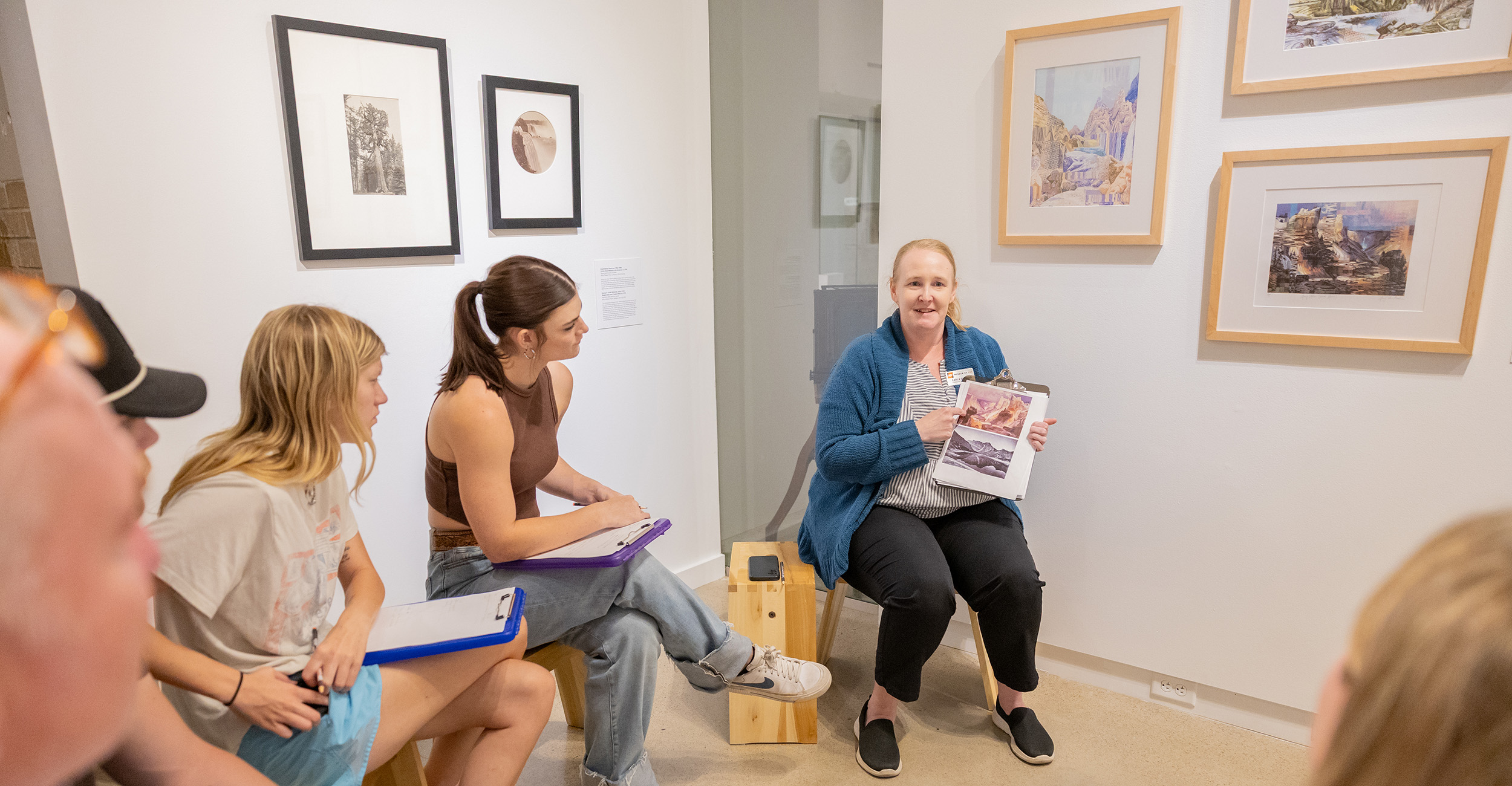 Group of people seated in a small gallery space with framed artwork on the walls. One person is holding up a book or catalog and appears to be leading a discussion, while others sit on benches with clipboards or notebooks.