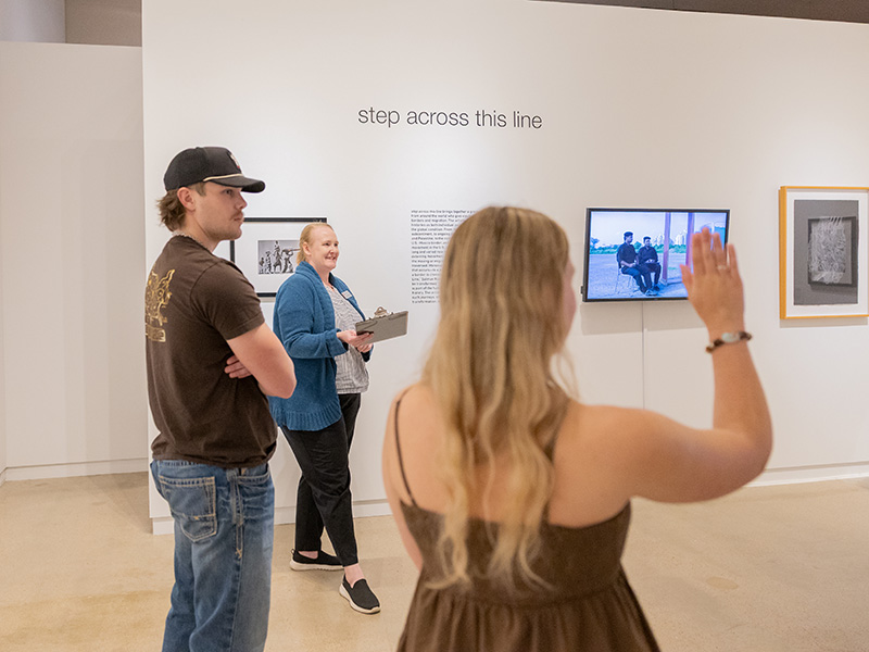 Three people standing in an art gallery near a wall with text reading “step across this line.” One person holds a clipboard, another wears a dark T-shirt and cap, and a third person in the foreground raises a hand toward a video screen mounted on the wall. Framed artwork and a monitor displaying an image are visible.