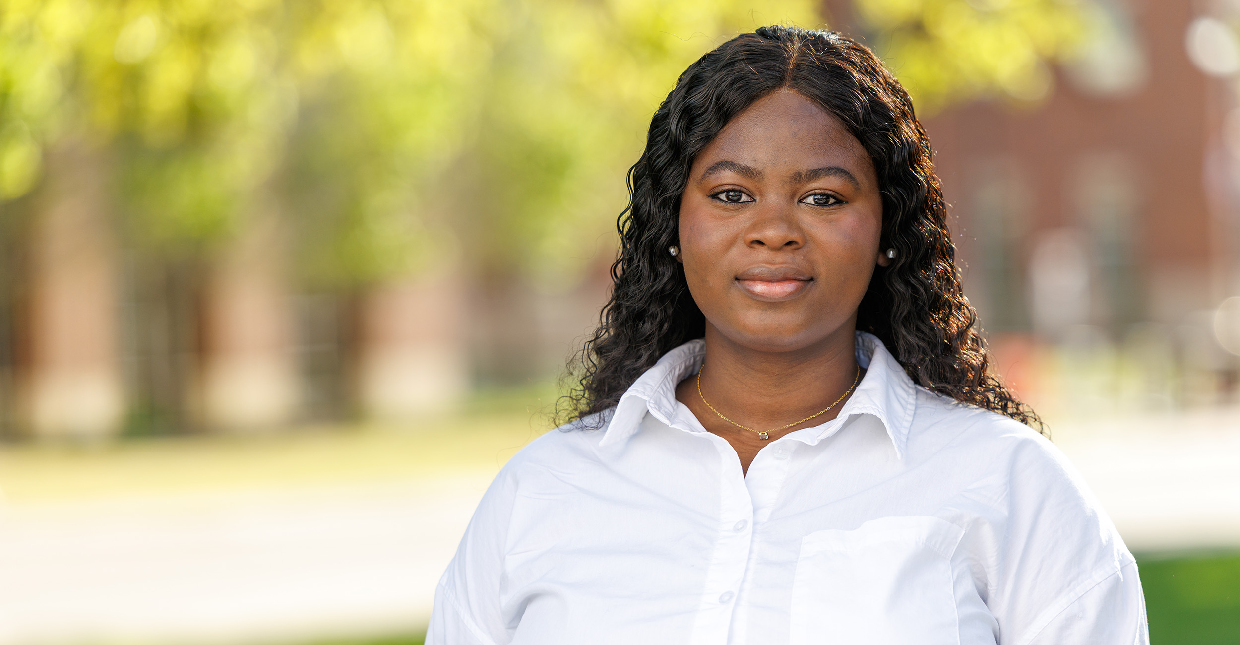 Portrait of a person wearing a white button‑down shirt standing outdoors on a green, tree‑lined campus with a building softly blurred in the background.