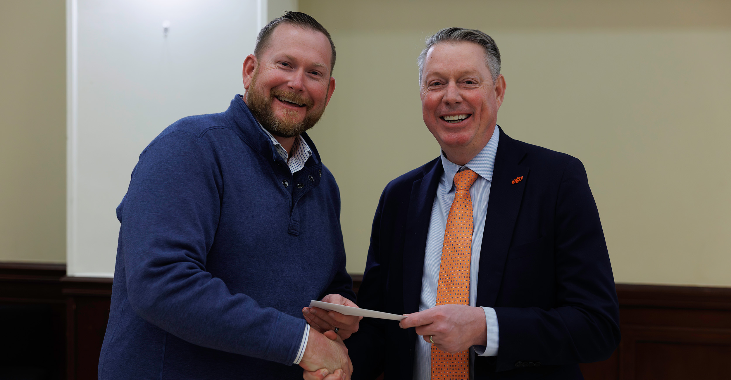 Chris Eck poses for a picture and smiles. He wears a blue sweater and is holding onto a clear plaque being presented to him by Dr. Jayson Lusk, vice president and dean of OSU Agriculture. Lusk wears a dark suit. He also poses and smiles for the picture.
