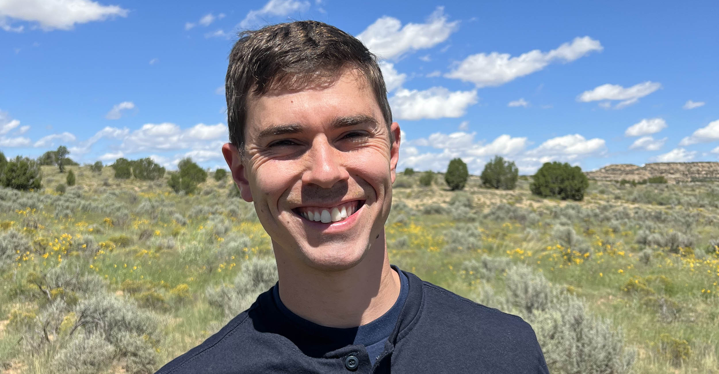 Nolan Craun stands in the sage rangeland of New Mexico with dry ground, green shrub plants and blue skies in the background. He is smiling, with brown hair and wearing a navy blue shirt.