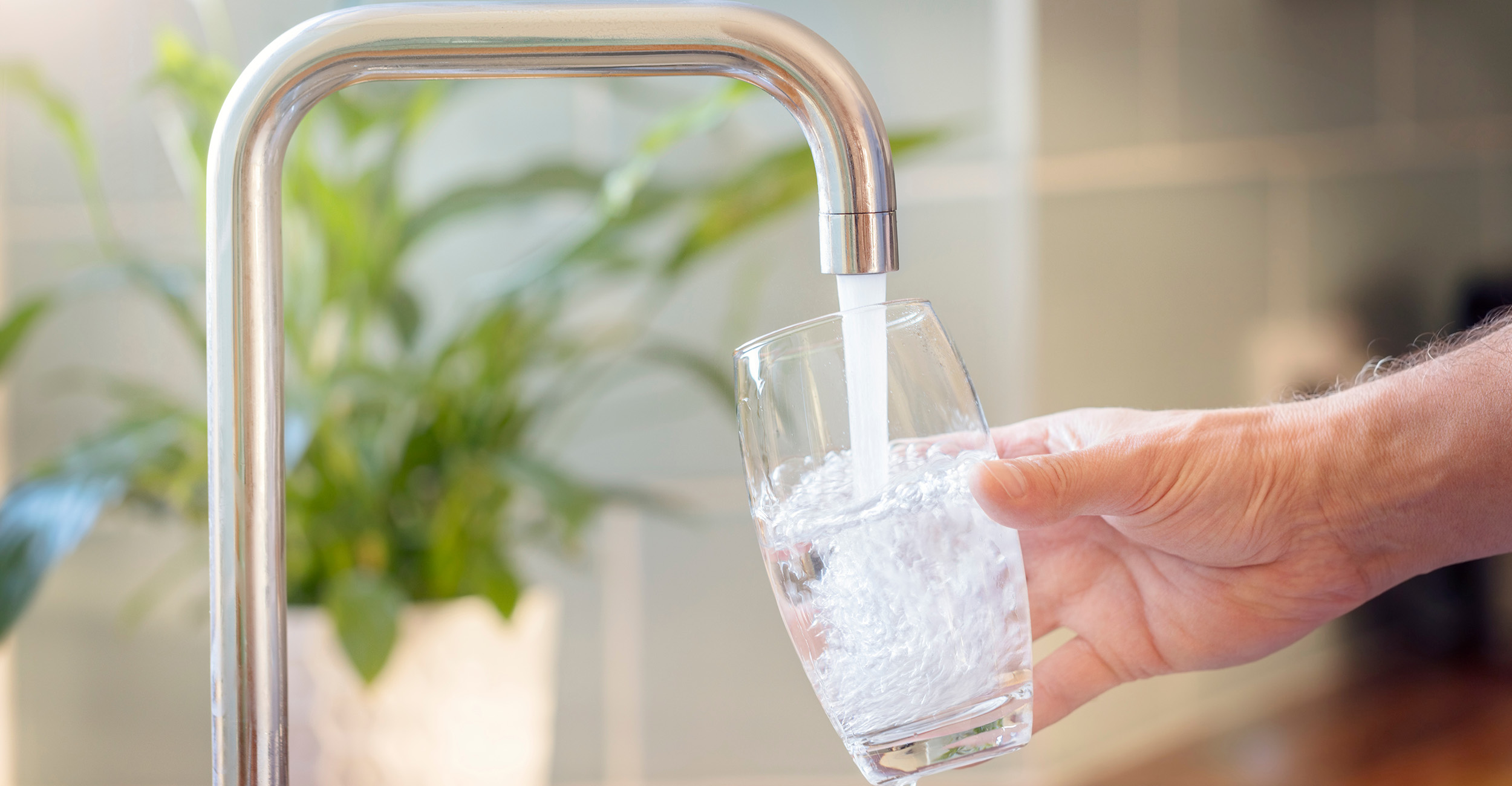 A hand holds a clear drinking glass under a modern kitchen faucet as fresh water pours in, illustrating access to clean, safe drinking water.
