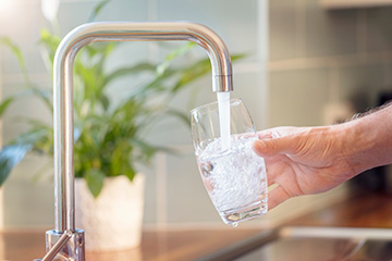 A hand holds a clear glass under a kitchen faucet as clean water flows into it, illustrating access to fresh drinking water in a home setting.