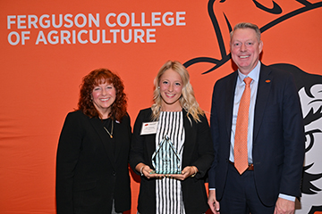Three people stand in front of an orange Ferguson College of Agriculture backdrop. The person in the center holds a glass award while posing for a photo with two individuals in business attire. The backdrop includes the text “Ferguson College of