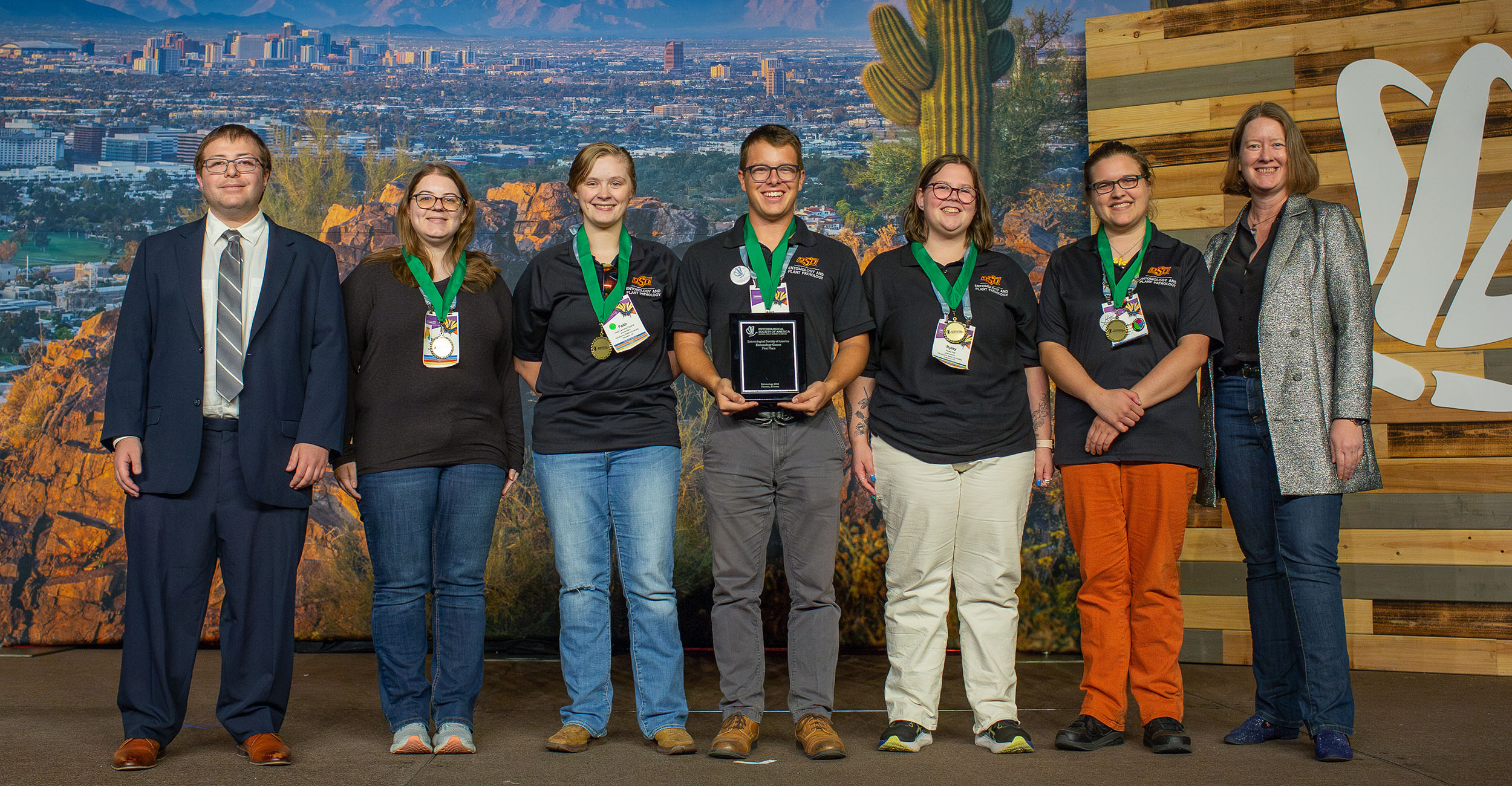 A group of seven men and women stand in front of a photo backdrop of a city skyline. The man on the far left is wearing a blue suit, tie and glasses. The woman on the far right is wearing jeans, a black blouse and a silver, glittery blazer. The male and female students in between are wearing black OSU polo shirts with jeans, grey pants or khaki pants.