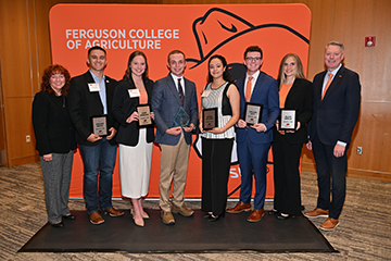 A group of eight people pose in front of a Ferguson College of Agriculture backdrop featuring the college name and a large Pistol Pete graphic. Five students hold award plaques, and one student in the center holds a glass recognition award. Two