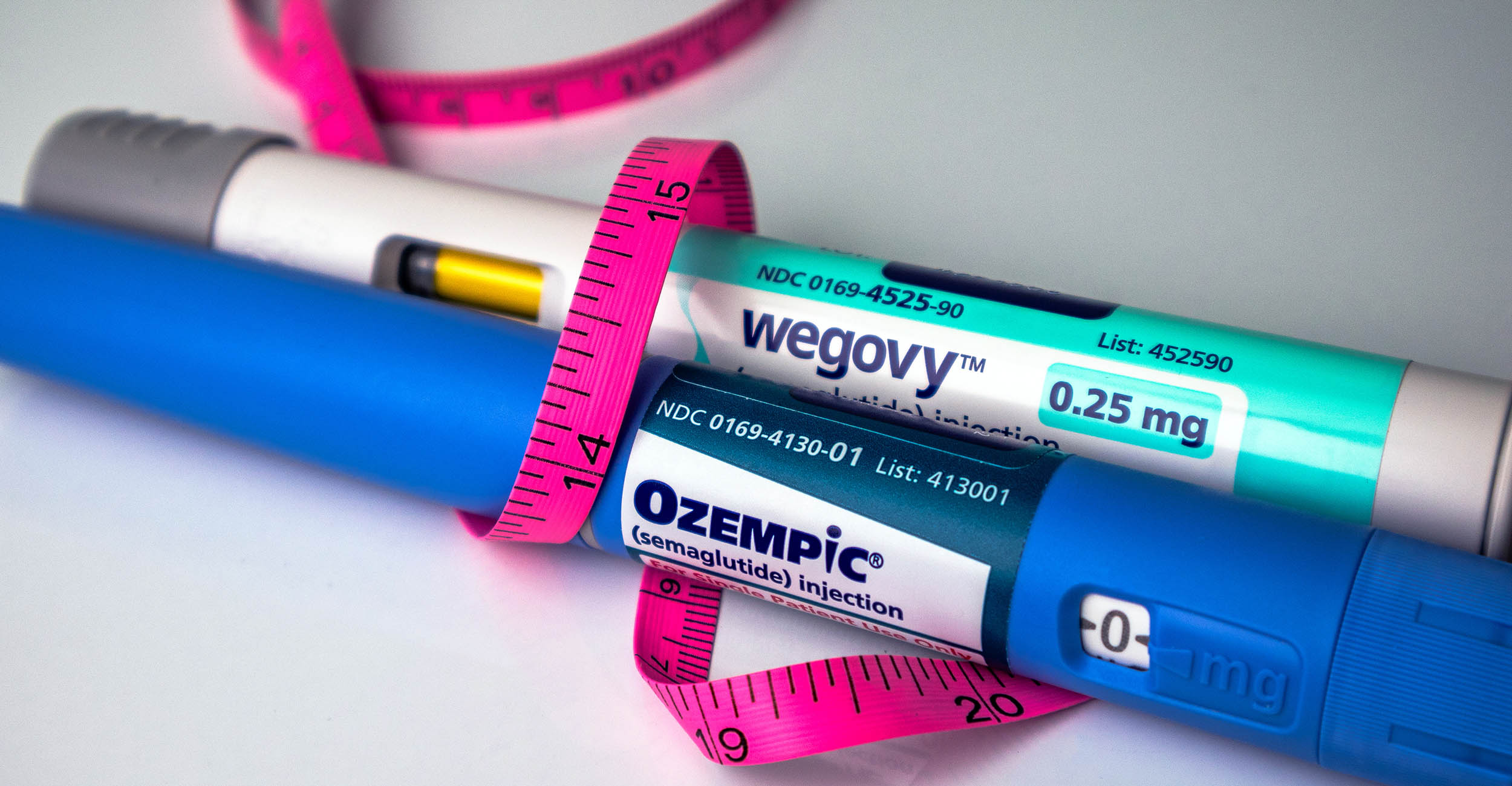 A stock photo of two tubes of medicine that humans inject with a needle. One tube is labeled Wegovy with a light green label. The other is labeled Ozempic with a blue label. A pink measuring tape is wrapped around both tubes.