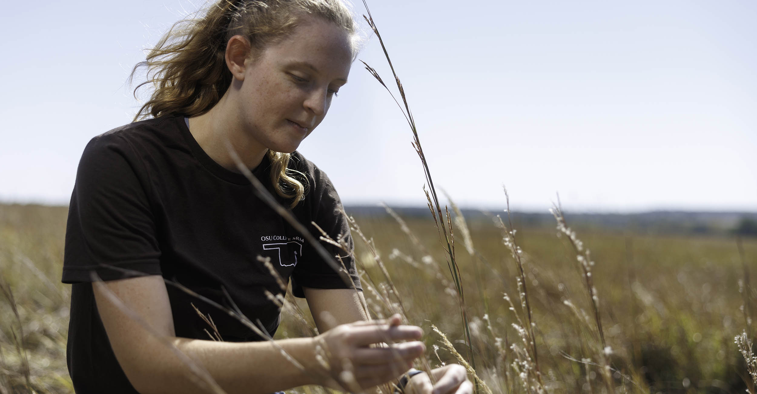 Kendall Christensen bends down in a field of golden brown native grasses. She is wearing a black T-shirt, jeans and her hair is in a ponytail.