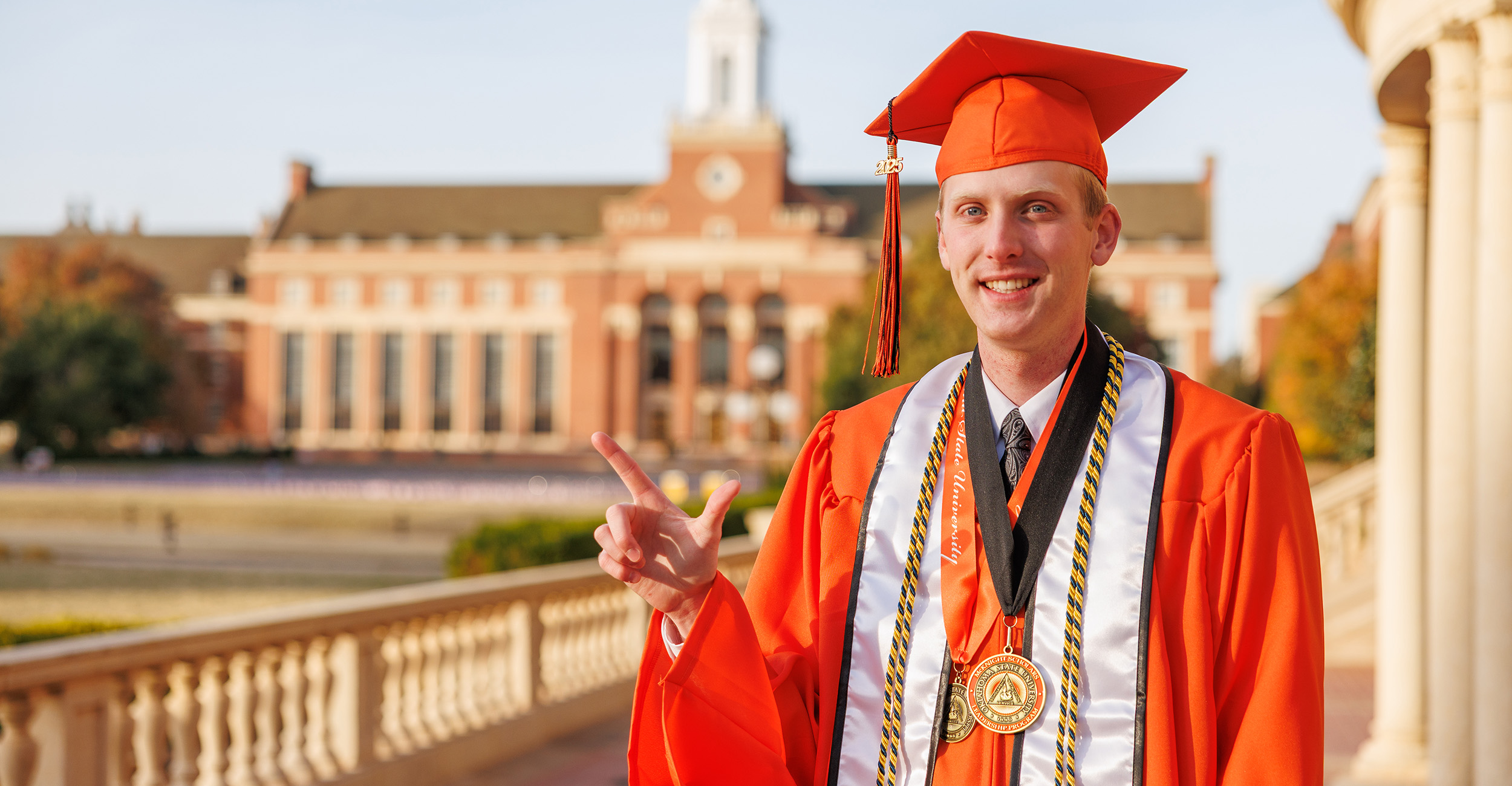 Cody Loganbill stands in front of the OSU Edmon Low Library, wearing an orange cap gown.