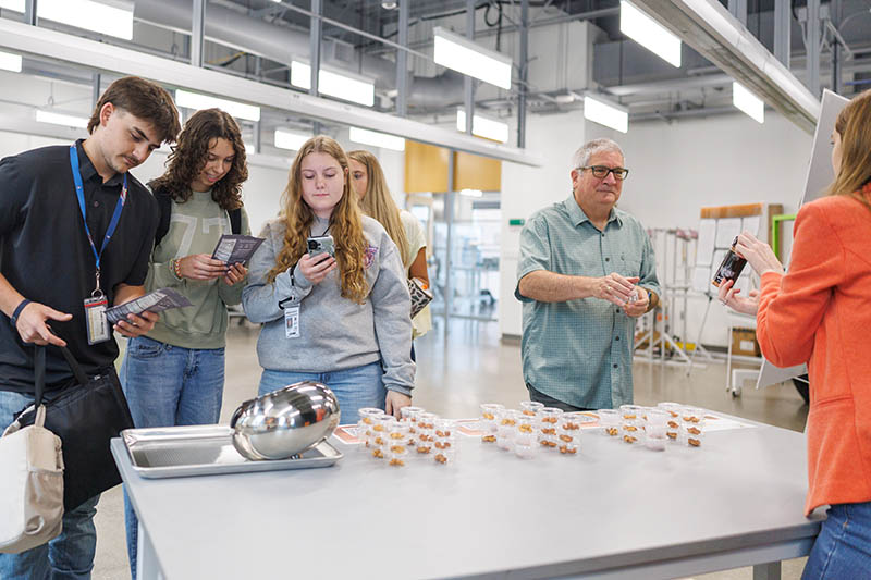 A group of students gathers around a table to sample and evaluate small food portions while a presenter explains the items, illustrating a hands‑on food science or product testing activity in a laboratory or teaching space.