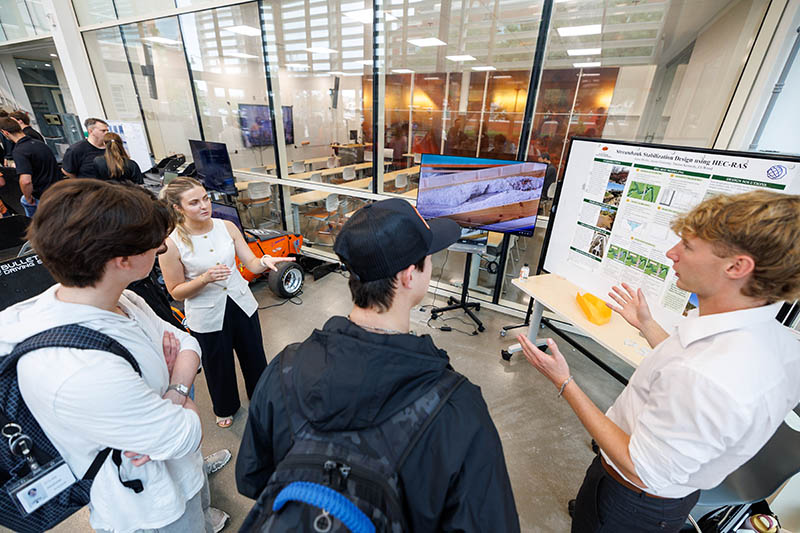 A group of students gathers around a display station as one student presents and explains a research poster and screen visualizations, illustrating an interactive academic demonstration or project showcase in a campus lab or innovation space.