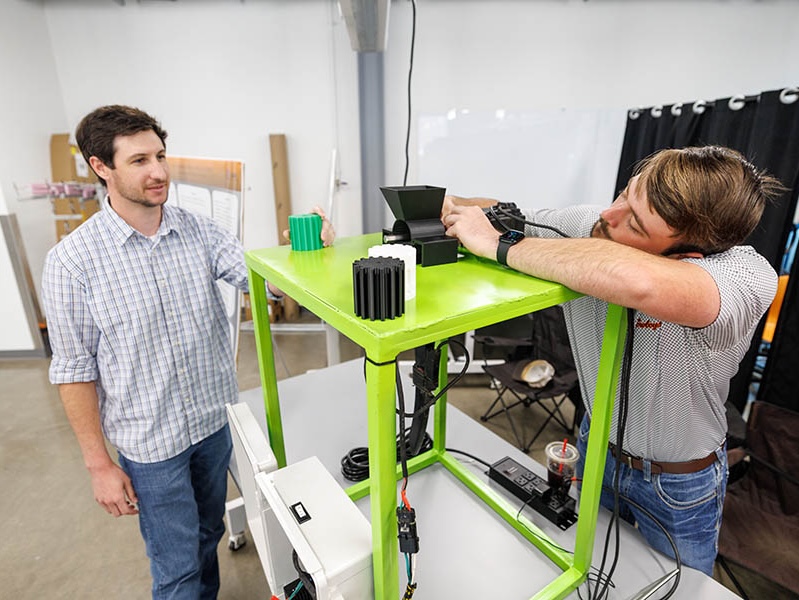 Two people work with a bright green prototype cart, adjusting components and equipment on top, illustrating hands‑on engineering testing or prototype development in a laboratory or workshop setting.