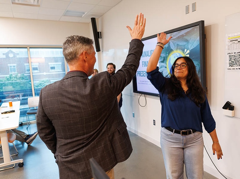 Two people stand facing each other in a classroom or innovation space, raising their hands for a high‑five in front of a presentation screen, illustrating collaboration and celebration during an academic or project presentation.