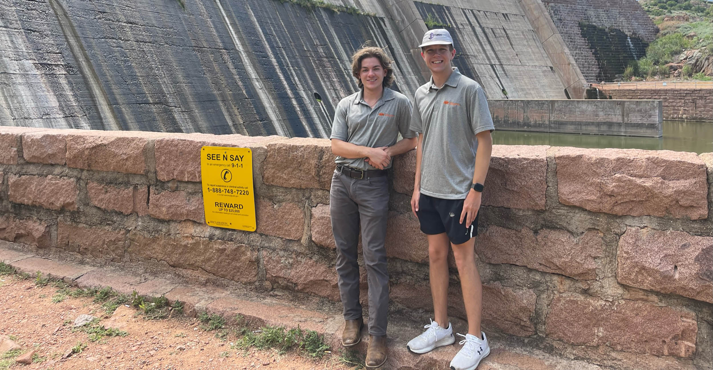 John Clemmons and Hoyt Nebgen stand in front of a dam water structure. John is in gray pants, brown boots and a gray polo shirt. Hoyt is wearing black shorts, white sneakers and a gray polo.