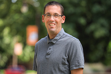 Scott Loss poses for a portrait image. He wears glasses, a gray polo shirt and is standing outside, with a tree in the background.
