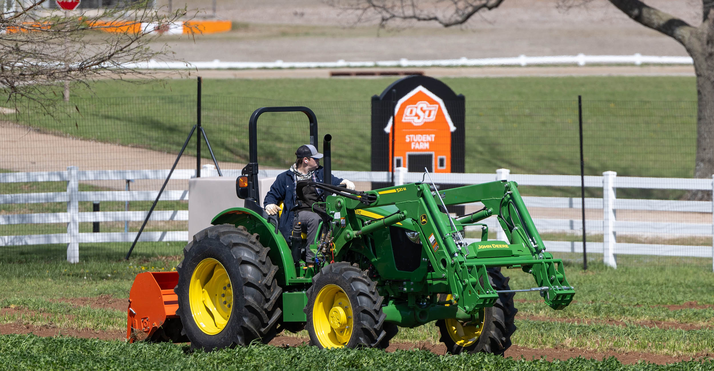 A young male adult drives a green John Deere tractor without a cab. The tractor is pulling a small orange disk. The student is wearing a blue coat and a black and gray hat.